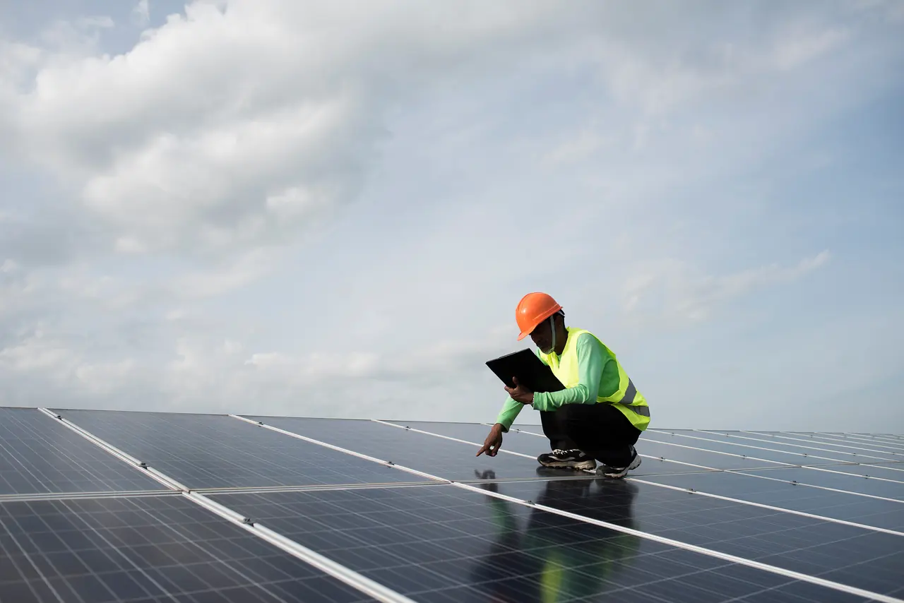 Technician in an orange hard hat and hi-vis vest crouching on solar panels with a tablet, pointing at a module